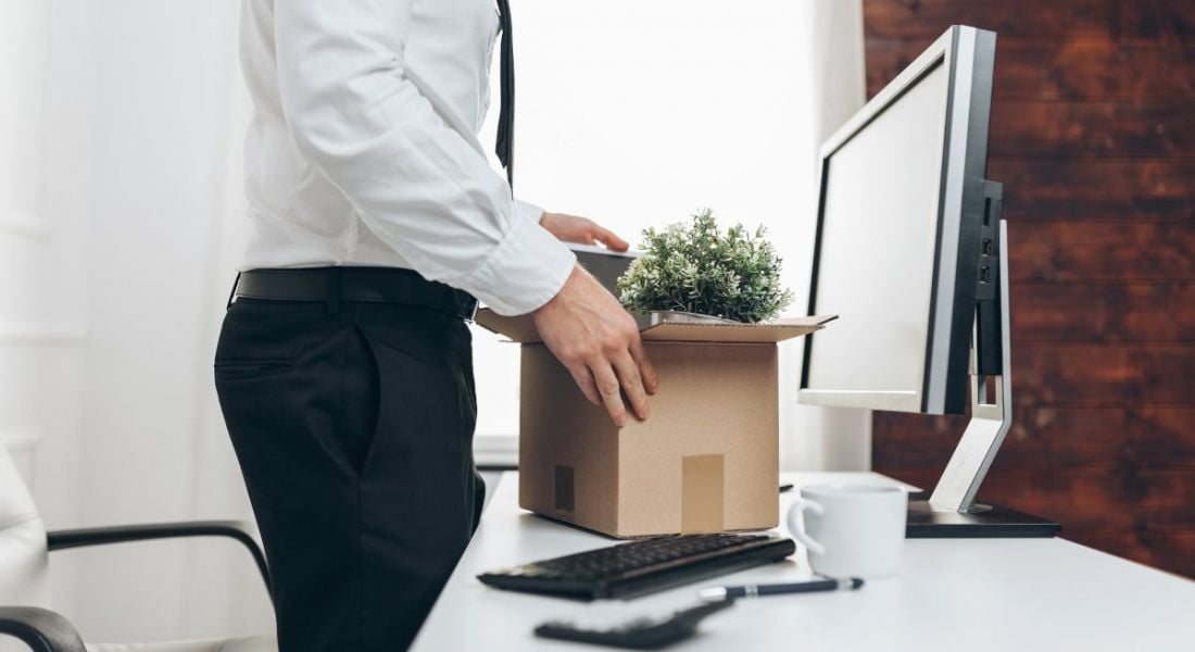 A man standing up at a desk with a cardboard box of items in it, symbolising redundancy.