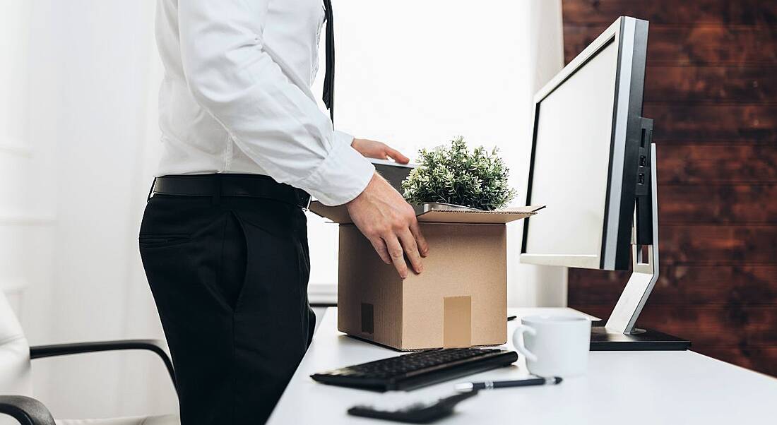 A man standing up at a desk with a cardboard box of items in it, symbolising redundancy.