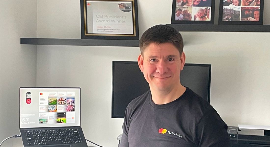 A man sits and smiles in a home office with a laptop, monitor and picture frames visible in the background.