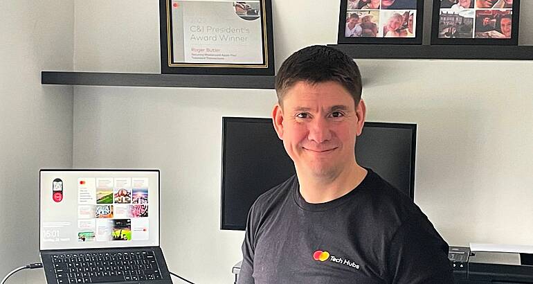 A man sits and smiles in a home office with a laptop, monitor and picture frames visible in the background.
