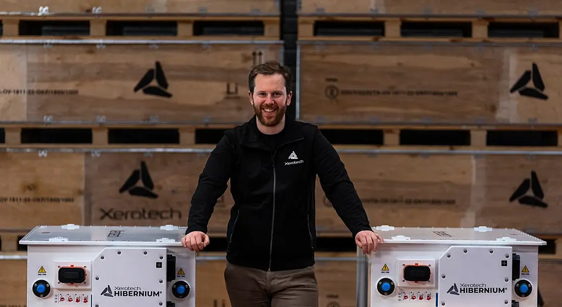 A man standing in the middle of two white battery packs with the Xerotech logo on them, with multiple brown boxes with the same logo behind him.