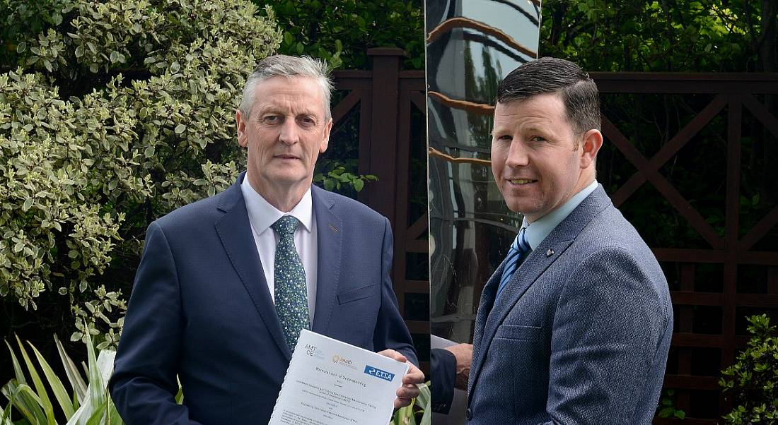 Two men standing outside in front of some leafy green trees and a statue. One man is holding a document.