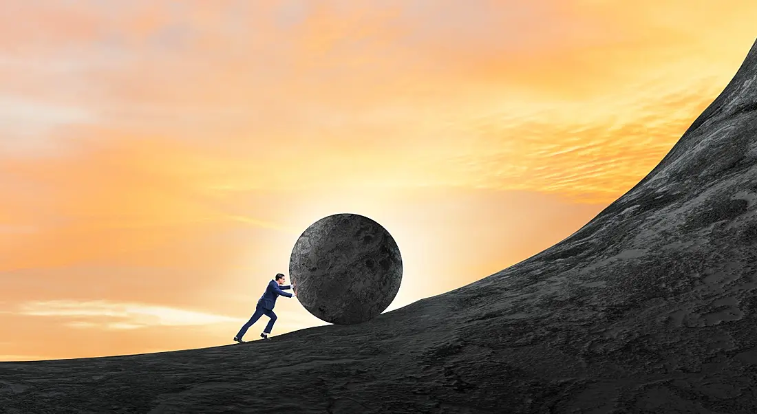 Struggling worker pushing a boulder up a very steep hill at sunset.