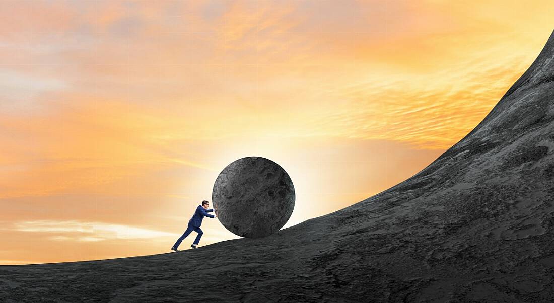 Struggling worker pushing a boulder up a very steep hill at sunset.