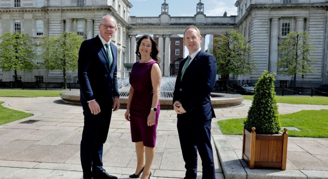 Two men and a woman standing in a row outside a majestic stone building on a sunny day.