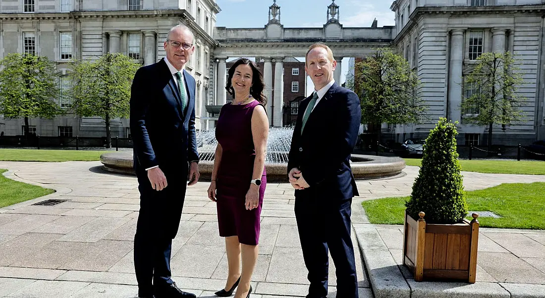 Two men and a woman standing in a row outside a majestic stone building on a sunny day.