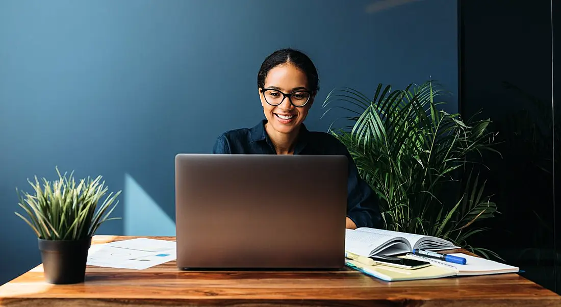 A woman smiling while working on a laptop. There are notes around her on the table.