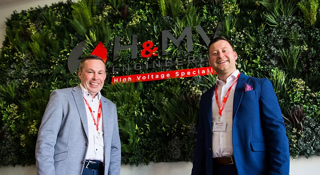 Two men in suits standing in front of a wall with the H&MV Engineering logo on it.