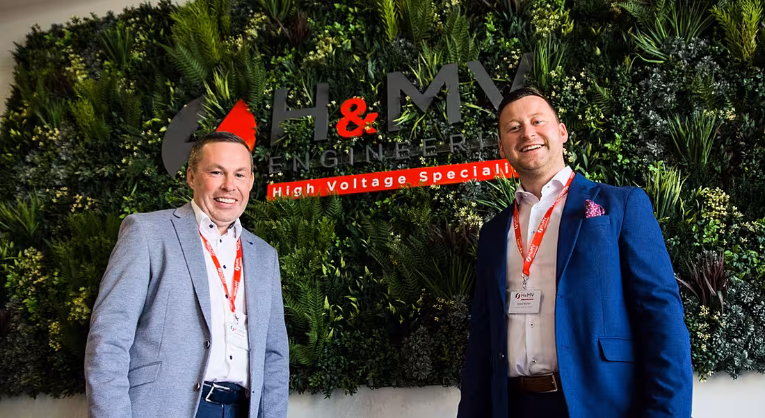 Two men in suits standing in front of a wall with the H&MV Engineering logo on it.