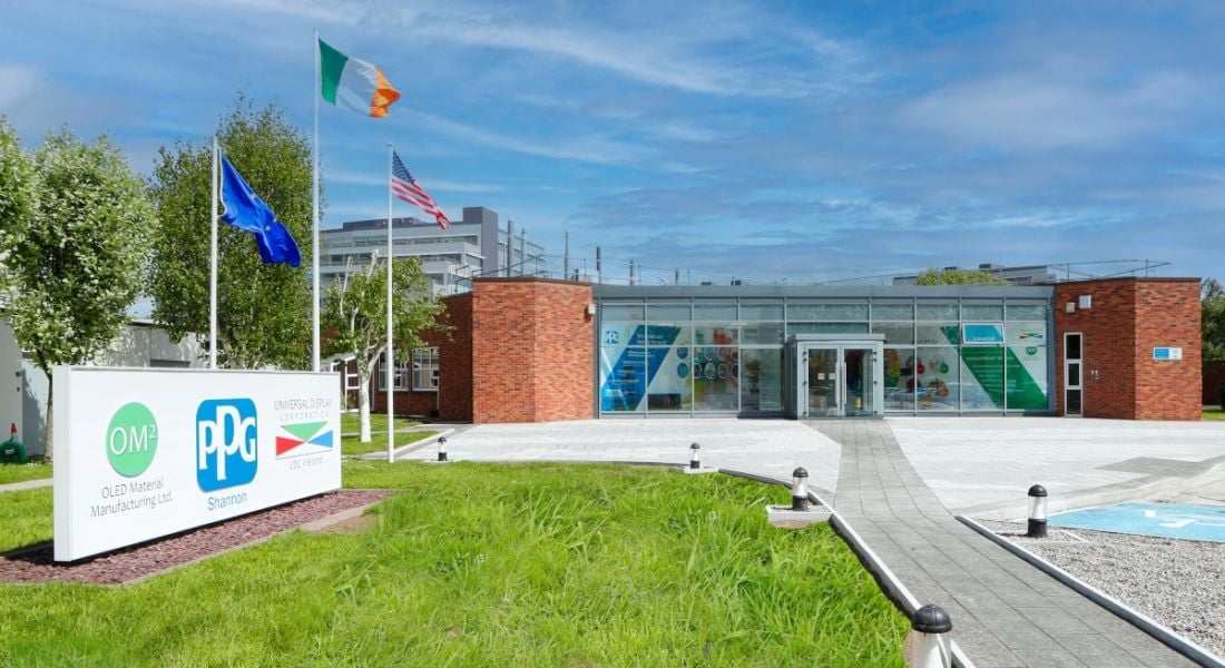 The exterior of a building with a signpost on some grass and the flags of the EU, Ireland and the US behind the sign.