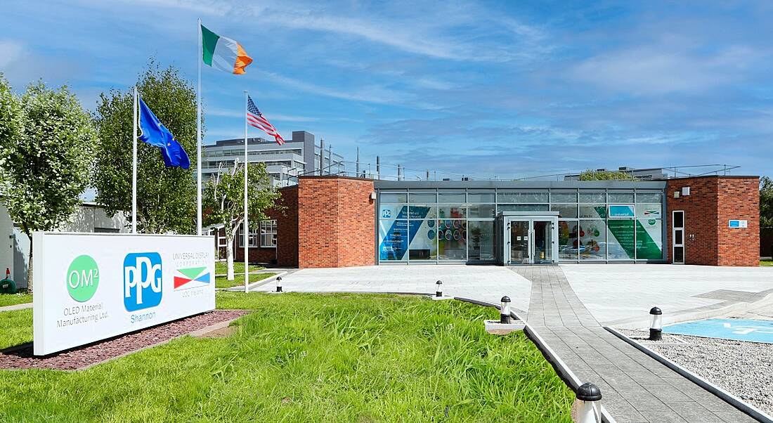 The exterior of a building with a signpost on some grass and the flags of the EU, Ireland and the US behind the sign.