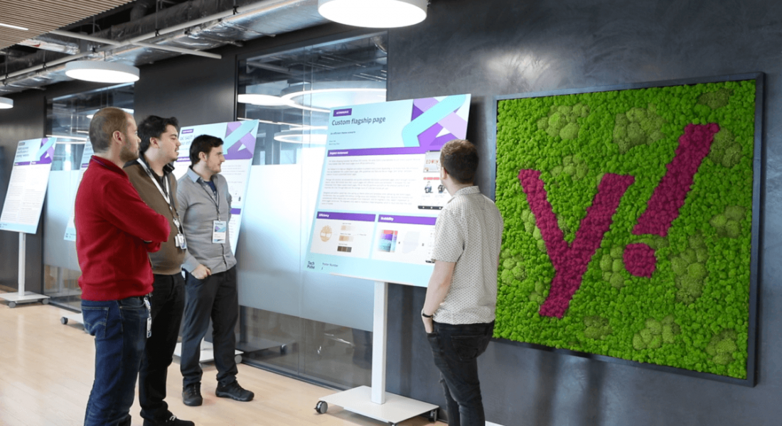 Four people stand around looking at an information board in an office. To the right of the board is a board depicting the letter 'Y' and an exclamation mark, representing Yahoo.
