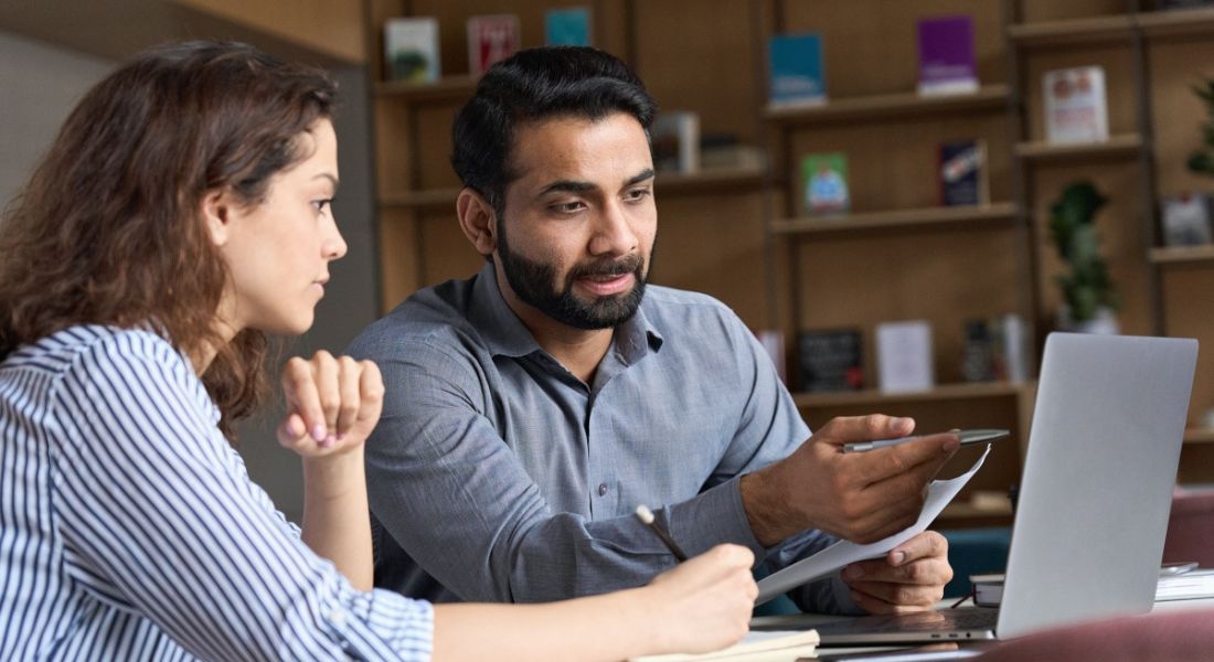 A young woman sitting next to a man showing a reverse mentorship as they work in front of a laptop.