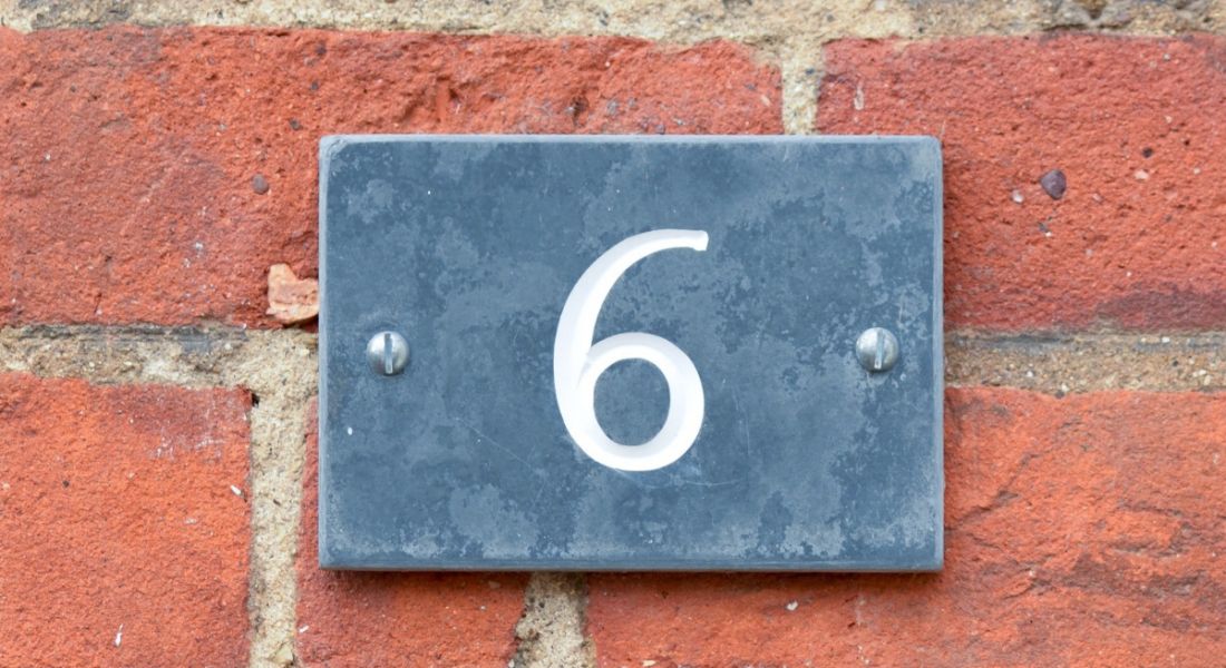 A grey sign with the number six nailed to a brick wall.