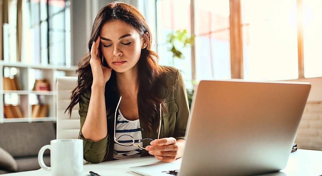 A stressed-looking woman sits at a desk with a computer. She has a hand up at her temple with her eyes closed, trying to combat stress.