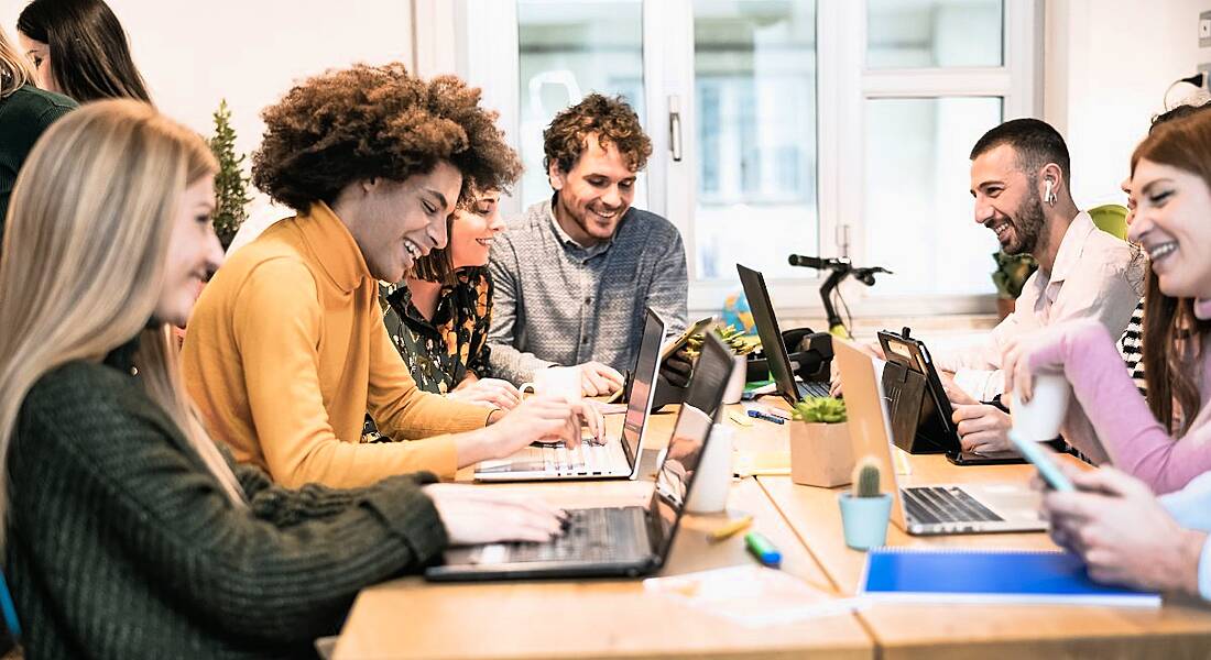 A group of early-stage talent sit around a boardroom table with laptops working and smiling.