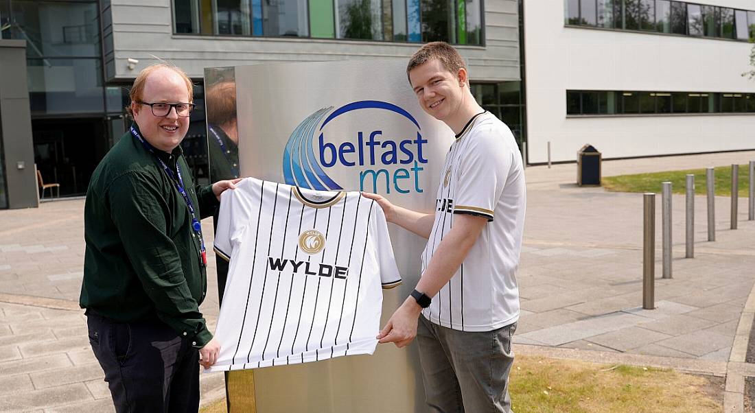 One man giving another man a jersey from Wylde outside a gray building with bollards visible in the background.