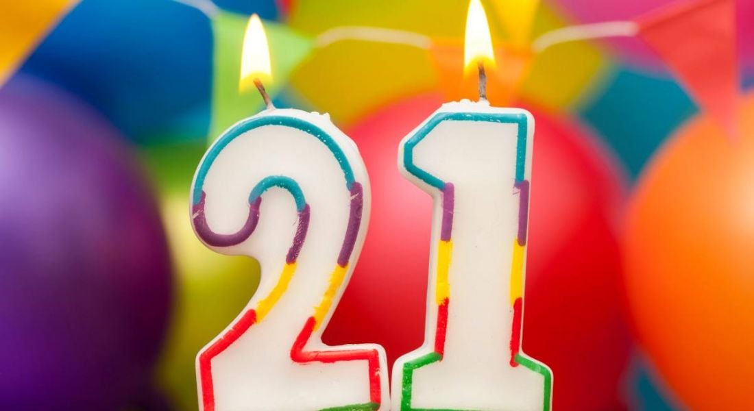 White candles shaped in the numbers 2 and 1 are lit and have multicoloured frames. There are ballons of different colours behind them.