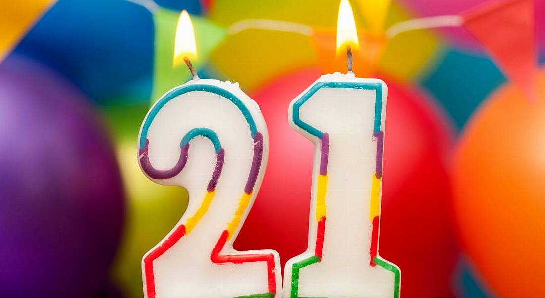 White candles shaped in the numbers 2 and 1 are lit and have multicoloured frames. There are ballons of different colours behind them.