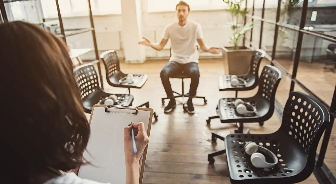 Man sitting in a chair at the end of a row of unoccupied chairs. A woman with a clipboard faces him and he gestures defensively at her.