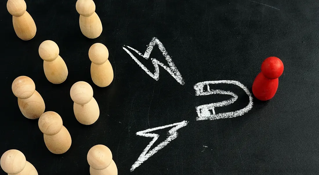 Talent attraction concept with wooden figurines facing one red figurine and chalk markings showing a drawing of a magnet and lightning symbols.