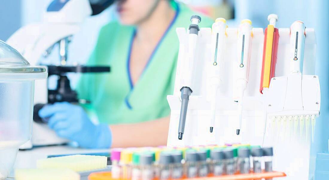 Woman working in a medtech environment lab looking at samples through a microscope. There are vials of liquid samples stored beside her.