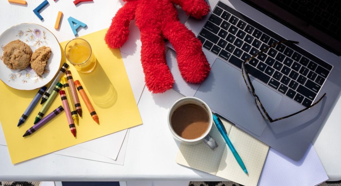 Working from home as a parent with kids concept. A red teddy bear and a full coffee mug and a pair of glasses on a laptop. There is a sheet of coloured paper with crayons and a plate of biscuits and glass of juice also.