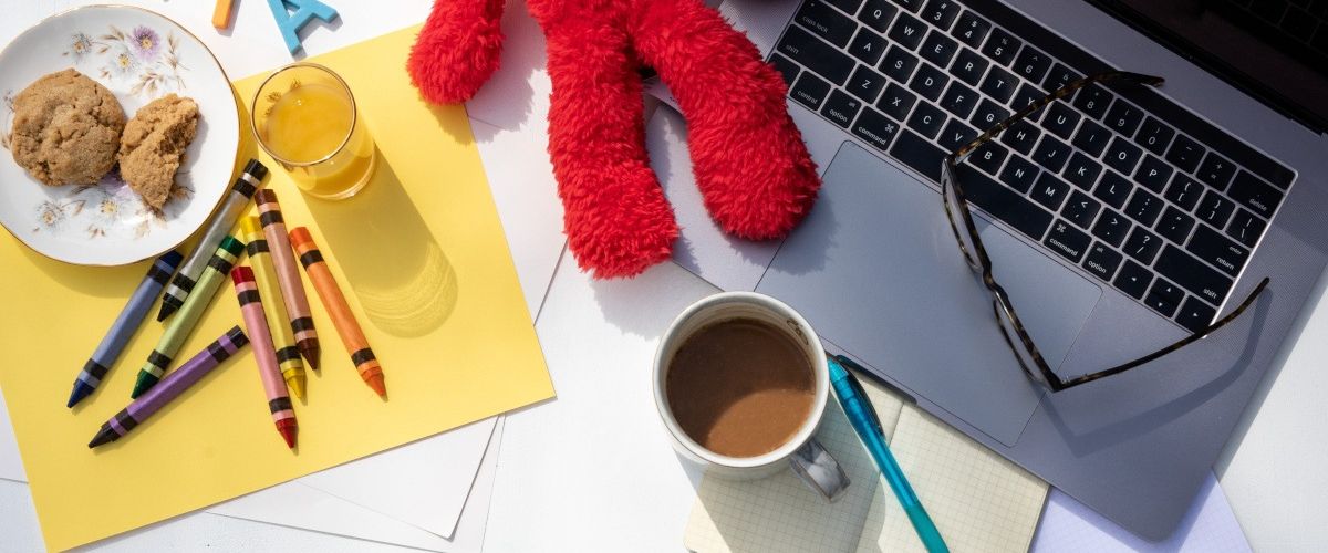 Working from home as a parent with kids concept. A red teddy bear and a full coffee mug and a pair of glasses on a laptop. There is a sheet of coloured paper with crayons and a plate of biscuits and glass of juice also.