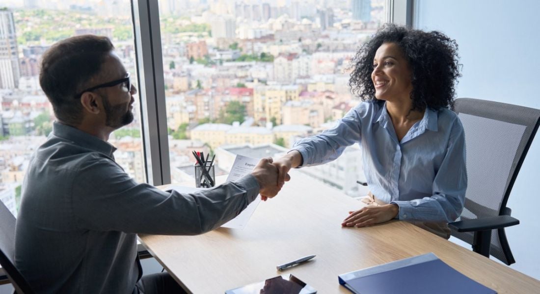 Man and a woman shaking hands across a table in a modern office.