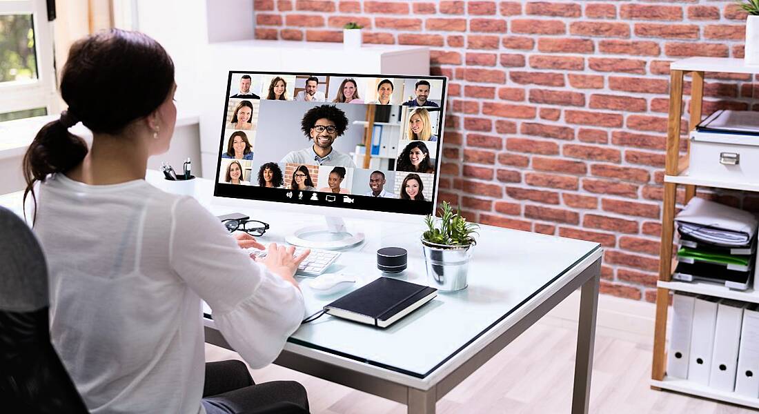 Woman working remotely in a modern office with red brick walls. She is attending a webinar on her computer as there are faces visible on her monitor and she has a notebook and pen on her desk as if she is taking notes.