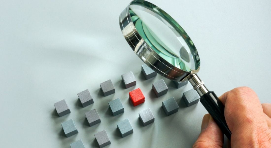 Person's hand holding a magnifying glass over some grey cubes with one red cube in the centre.
