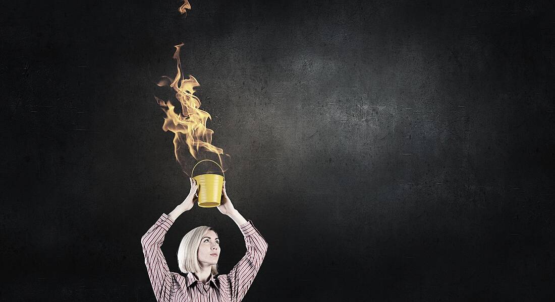 Young woman holding a bucket of fire above her head and looking up at it. There is a grey background.