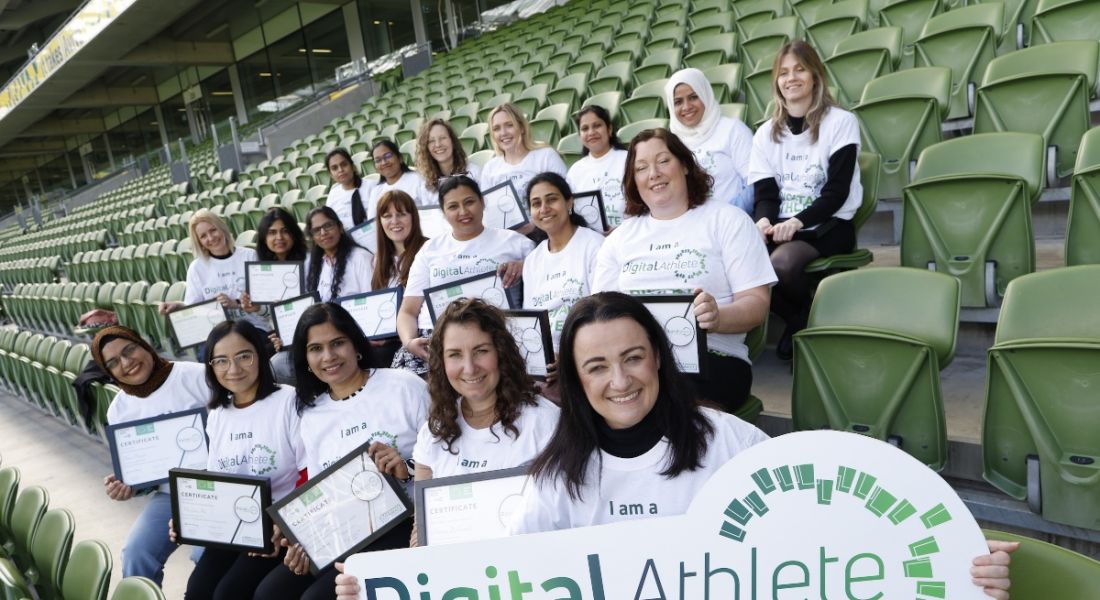A group of women sitting in the seats of a stadium wearing identical white t-shirts and smiling. The women in the front row of seats are holding a banner for the Digital Athlete programme.