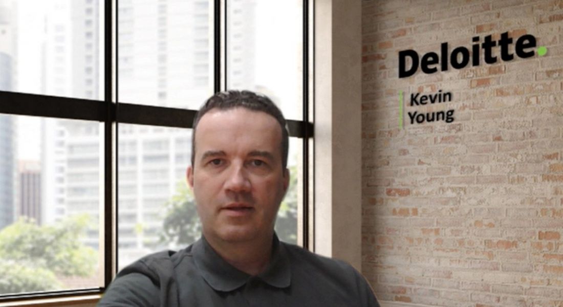 A man sits in an office with a window and wall behind him. The wall has Deloitte written on it as well as the man's name, Kevin Young.
