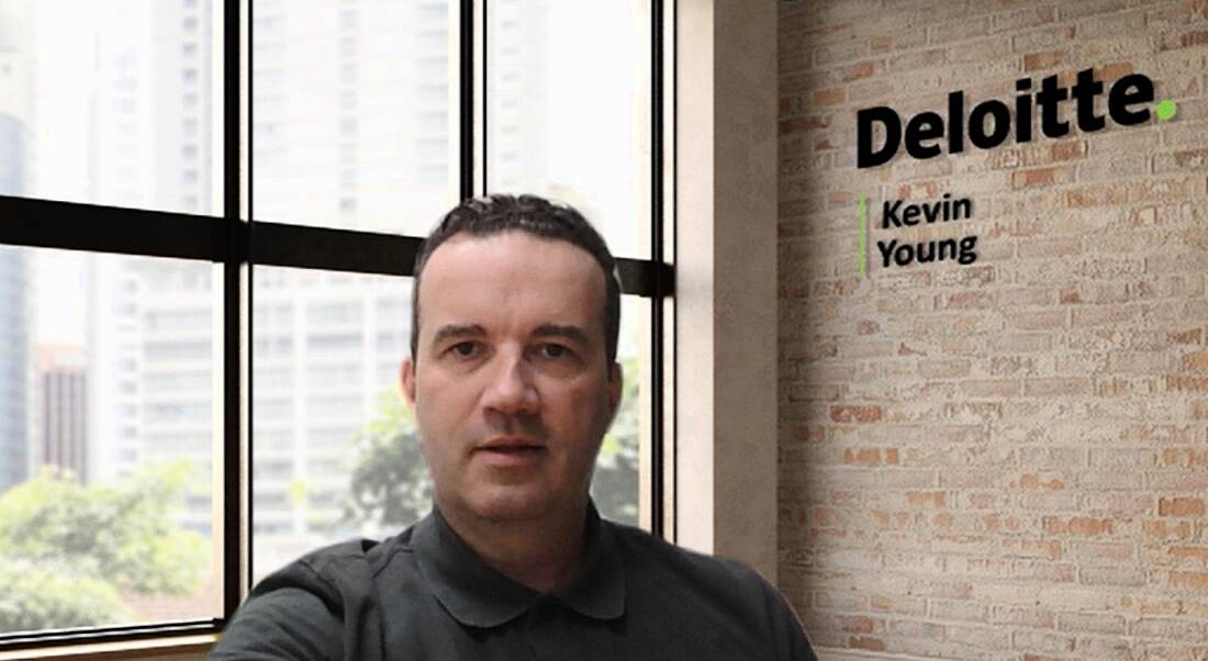 A man sits in an office with a window and wall behind him. The wall has Deloitte written on it as well as the man's name, Kevin Young.