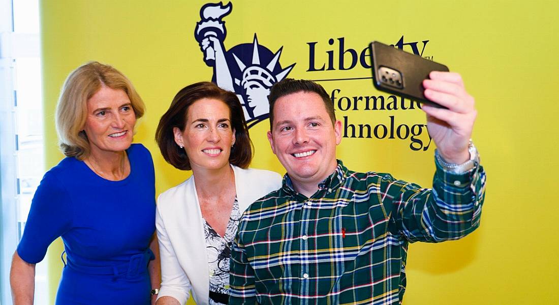 Two women and a man stand together with the man taking a photo of all three of them on his phone. They are standing in front of a wall that says Liberty IT.