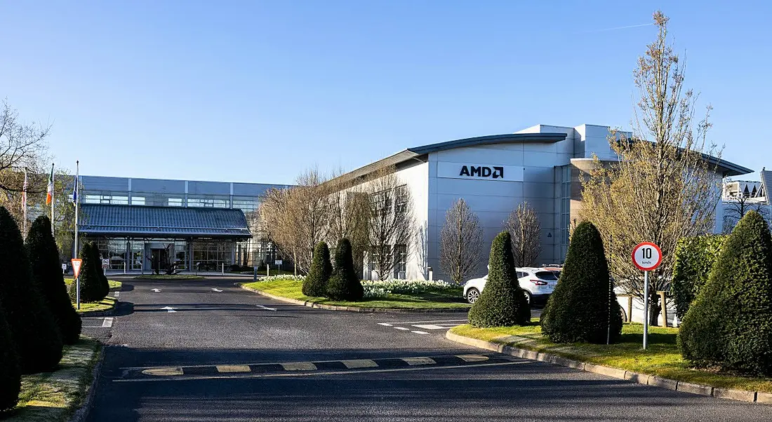 A large industrial building that is the AMD facility in Dublin. The front of the building and a large driveway with some trees and a blue sky are visible.