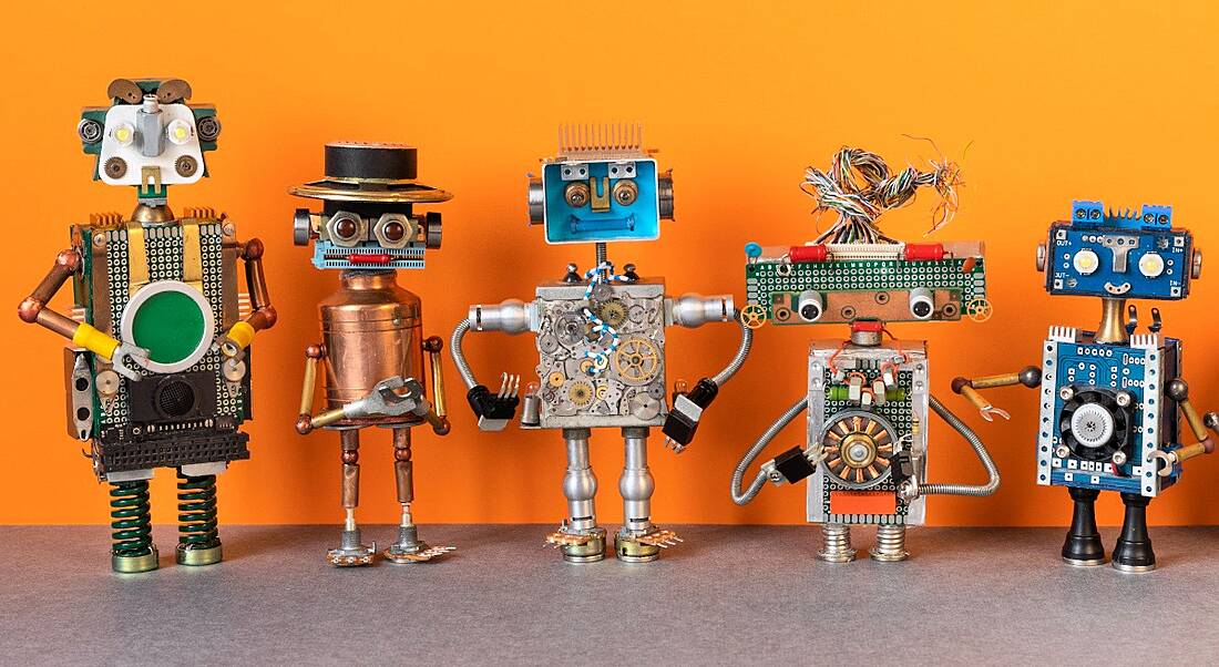 Five robots made from scrap parts of machines are set up in a line against an orange backdrop.