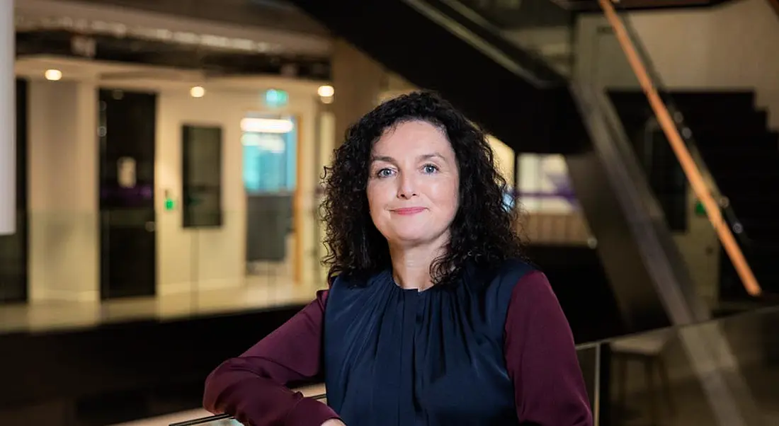 A woman leaning on a railing inside a building, with stairs and a door in the background. She is Sharon McCooey of LinkedIn Ireland.