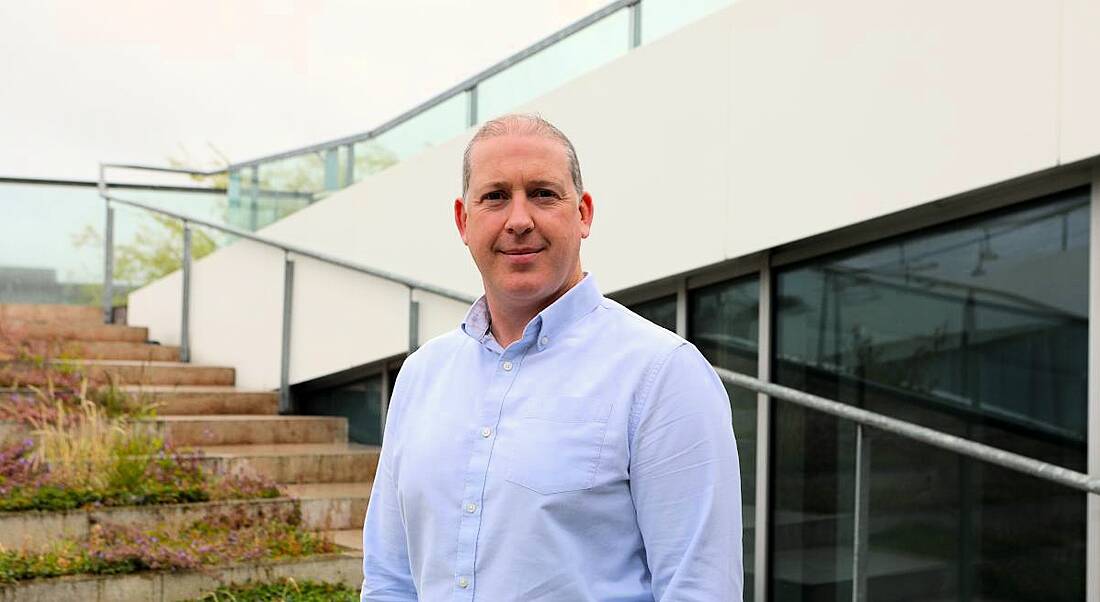 A man smiles at the camera wearing a light blue shirt while standing outside in front of a set of steps. He is Barry O'Brien, a network engineer at Aurora Telecom.