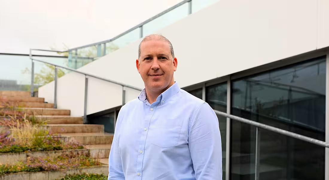 A man smiles at the camera wearing a light blue shirt while standing outside in front of a set of steps. He is Barry O'Brien, a network engineer at Aurora Telecom.