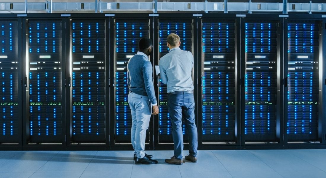 Two men standing facing a row of servers in a data centre.