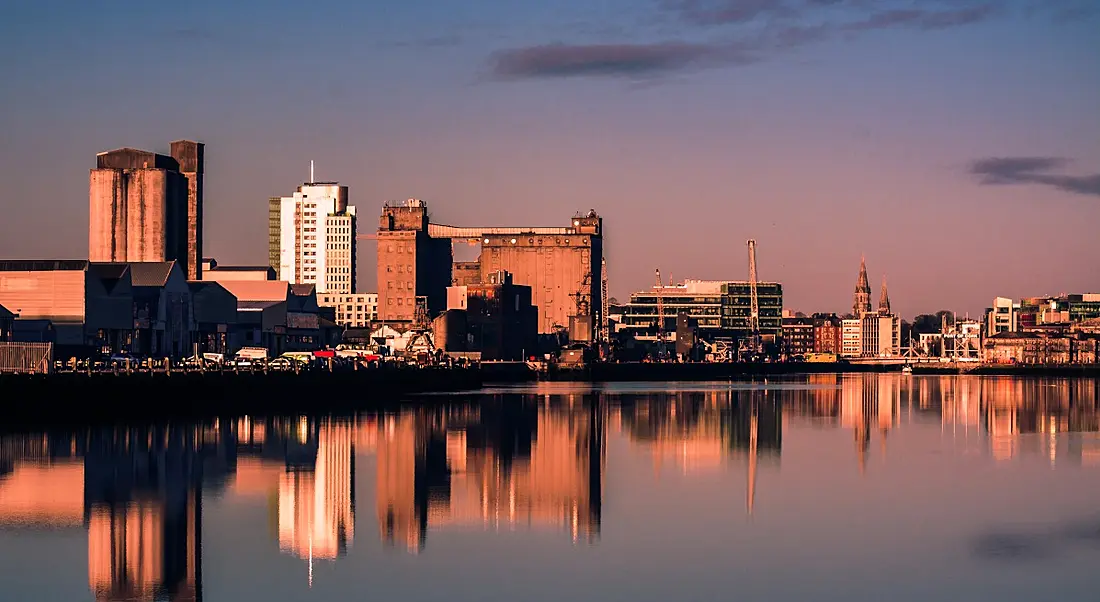 Buildings of Cork city, with reflections of the buildings visible on a river, with a sunset sky in the background.