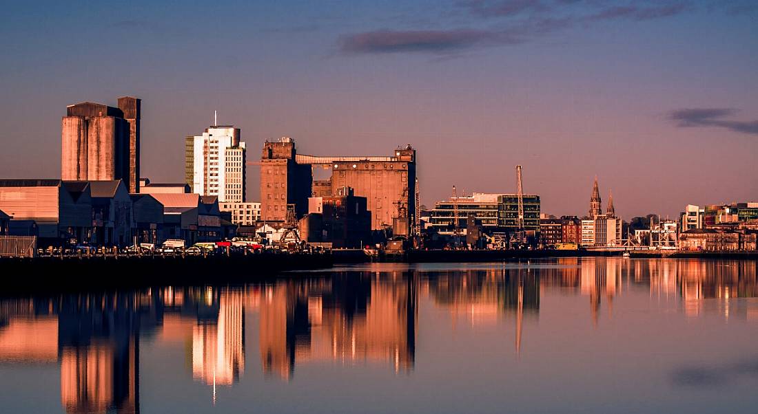 Buildings of Cork city, with reflections of the buildings visible on a river, with a sunset sky in the background.