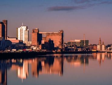 Buildings of Cork city, with reflections of the buildings visible on a river, with a sunset sky in the background.