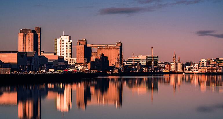 Buildings of Cork city, with reflections of the buildings visible on a river, with a sunset sky in the background.
