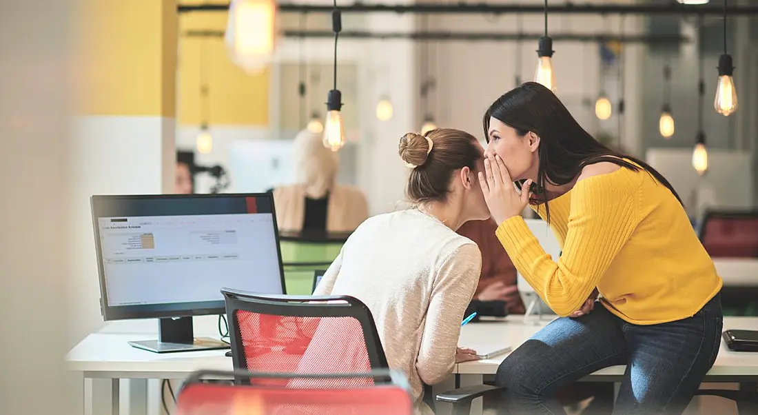 A woman sitting at her computer while a co-worker tells her a secret whispering in her ear.