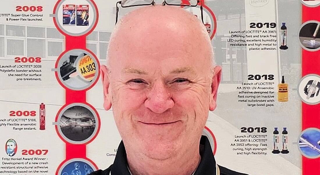 A man smiles while standing in front of an information board. He is Barry Burns, a technical lead at Henkel.