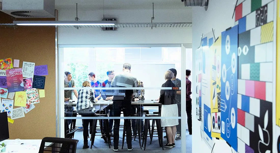 The Intercom office with posters and drawings on the walls. At the back a group of employees work in a meeting room behind a glass door.