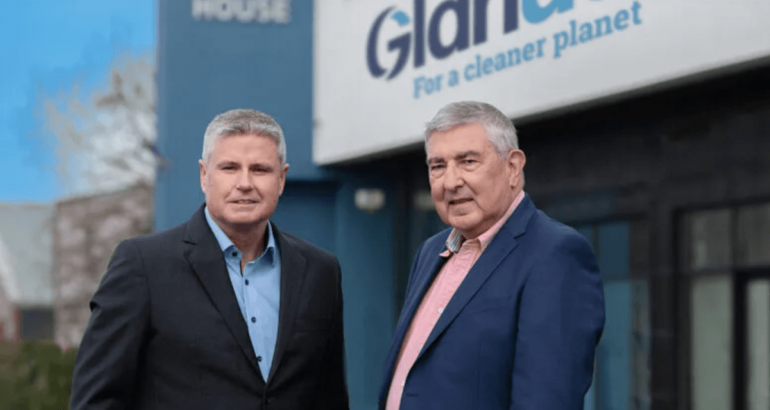 Two men in suits standing together in front of a building that has the Glanua logo on it.